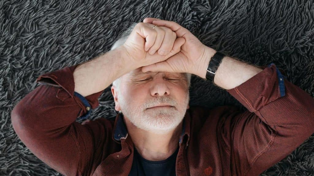 An older man lying on a carpet with his hands over his face.