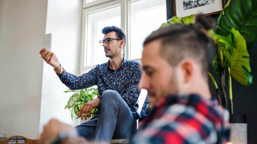 A man sits by a window holding a tablet, with another person blurred in front.
