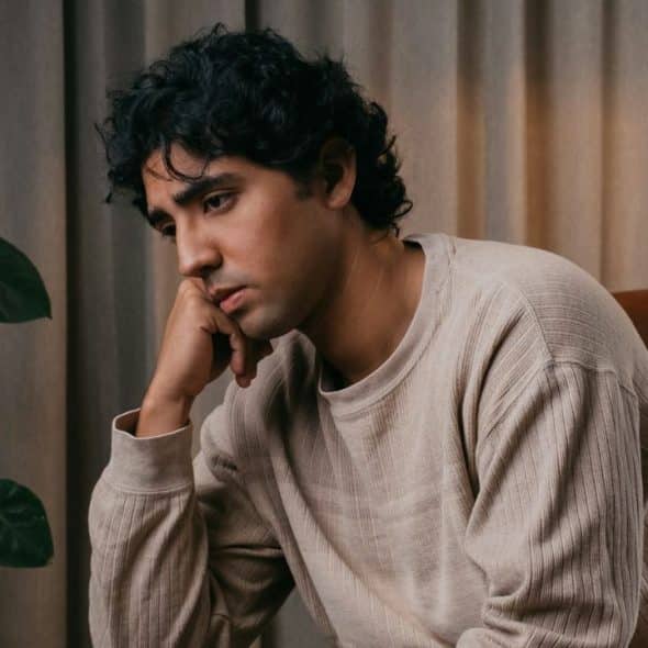 A thoughtful man sits indoors, resting his chin on his hand next to a houseplant.