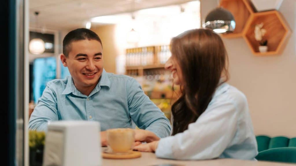 A man and woman are smiling and talking across a table in a cafe.