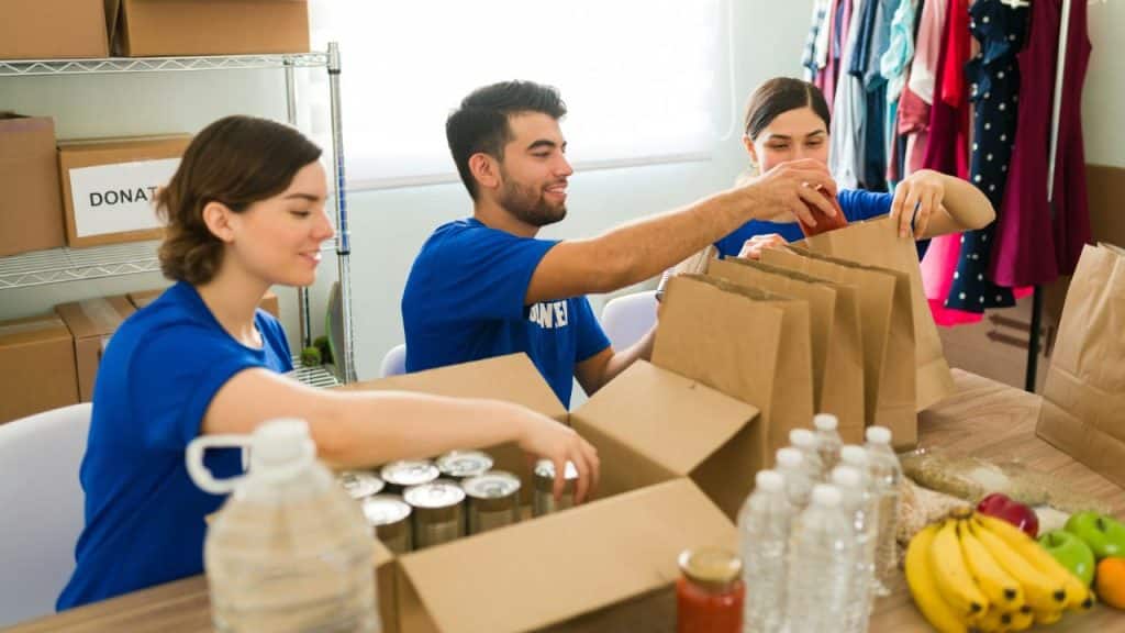 Three smiling volunteers in blue shirts are packing canned goods and fresh food into brown paper bags and boxes.