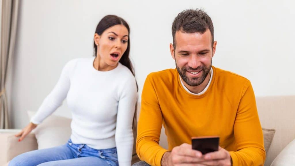 A man smiles at his phone while a surprised woman looks at him in the background.