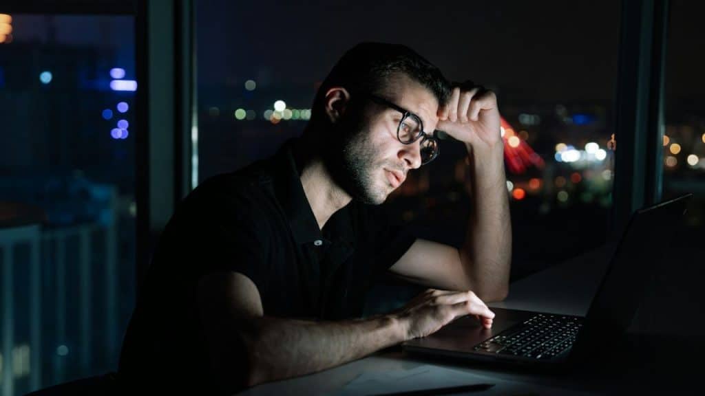 A man in glasses is working late on a laptop in a dark office, illuminated by the screen with city lights visible outside the window.