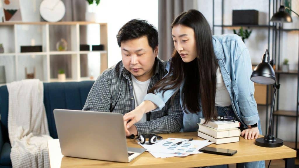 Two people are leaning over a laptop at a desk, looking at the screen together.