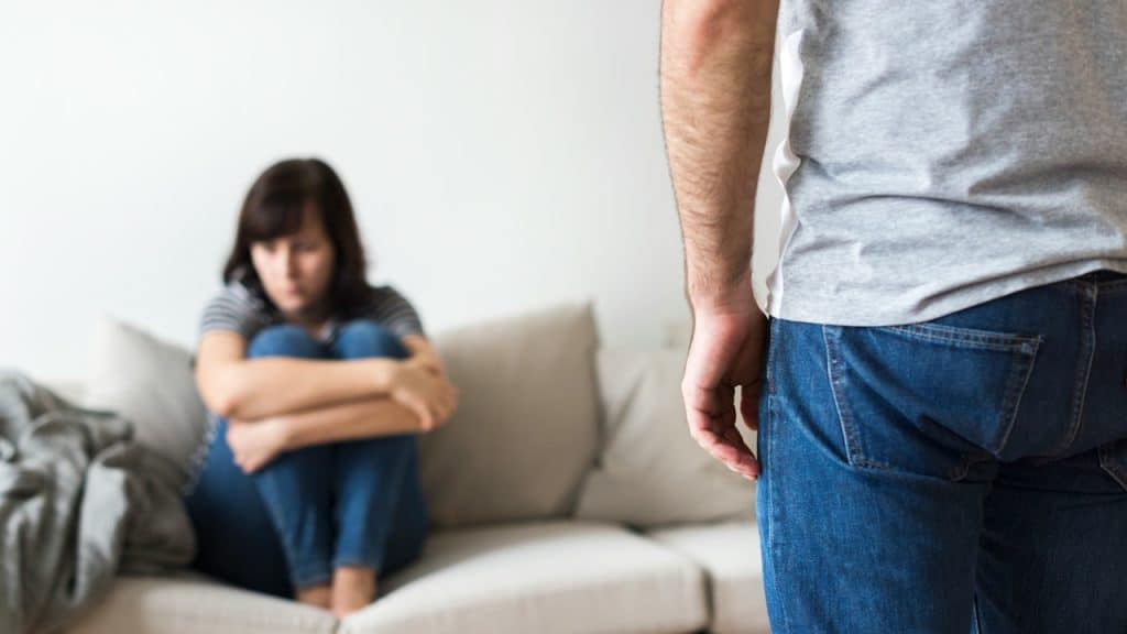 A man stands in the foreground with his back turned, while a distressed woman sits huddled on a couch in the background.