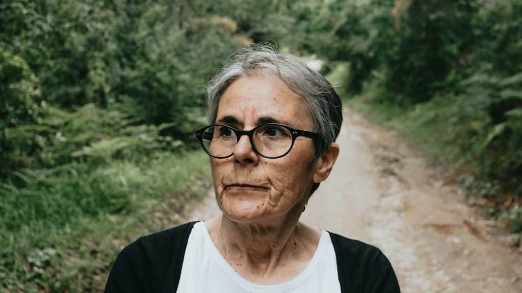 An older woman wearing glasses standing on a walking trek looking distraught.