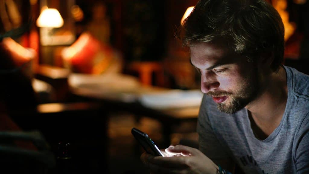A young, bearded man in a dark, softly lit interior looking down at his glowing smartphone held in his hands.