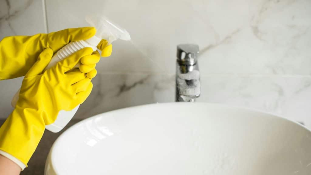 Hands in yellow gloves are spraying cleaner over a white bathroom sink.