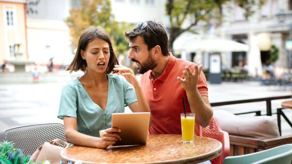 A frustrated man and an annoyed woman are arguing while sitting at an outdoor cafe.