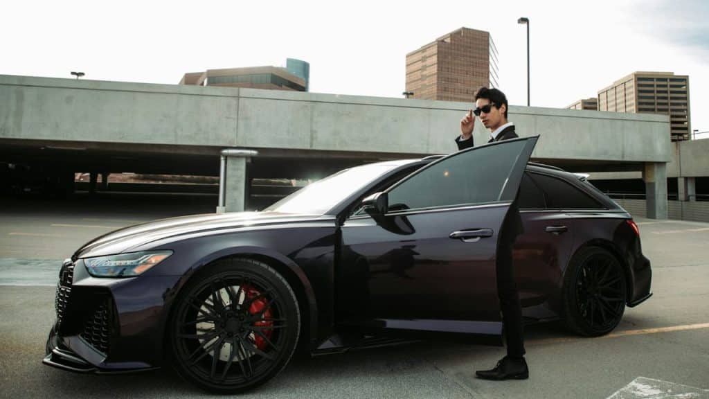Man in a dark suit and sunglasses standing by the open door of a sleek, dark sports car in a parking structure.