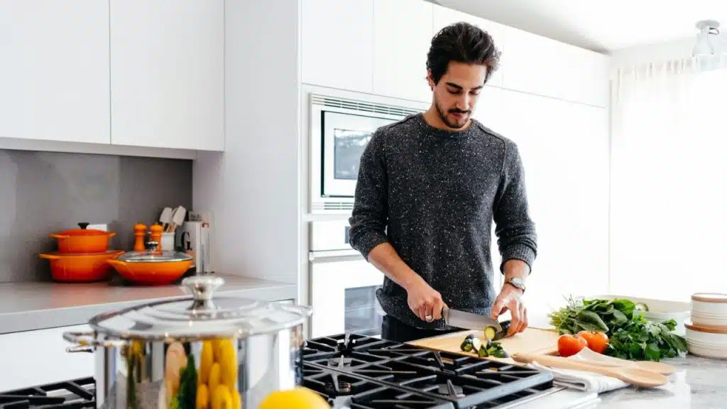Man in a speckled grey sweater slicing vegetables on a cutting board in a modern kitchen.