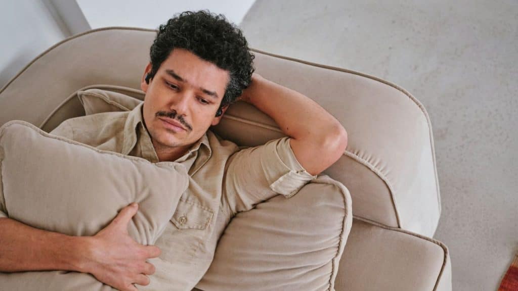 A man with a mustache lies on a beige couch with a pillow, wearing wireless earbuds.