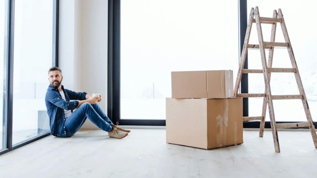 Man sitting on the floor by a large window with moving boxes and a ladder.