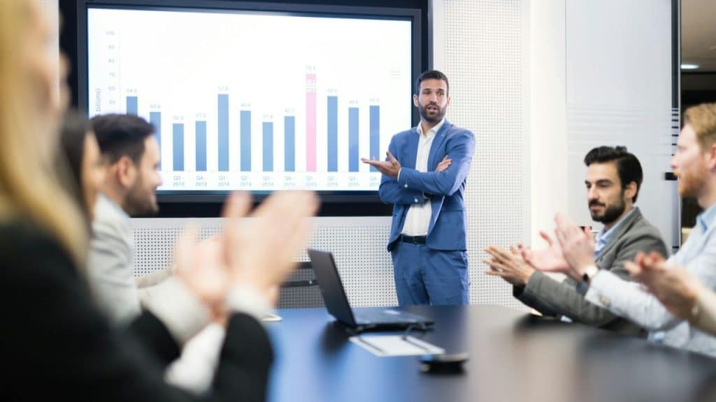 Man in a blue suit presenting a bar graph on a screen to a group of clapping colleagues.