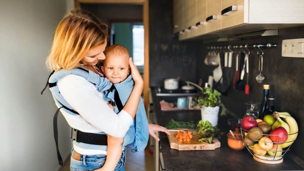 Woman holding a baby in a carrier and preparing vegetables on a cutting board in a kitchen.