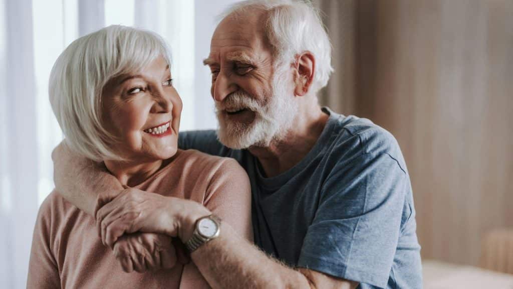 Smiling older couple with white hair hugging each other indoors.