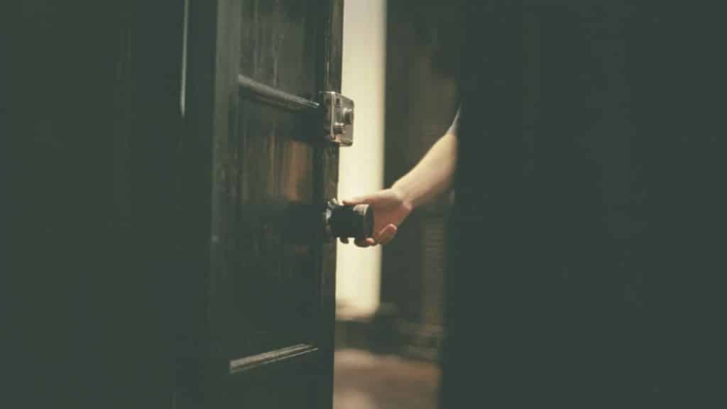 A hand opening a dark wooden door with a knob and latch visible.