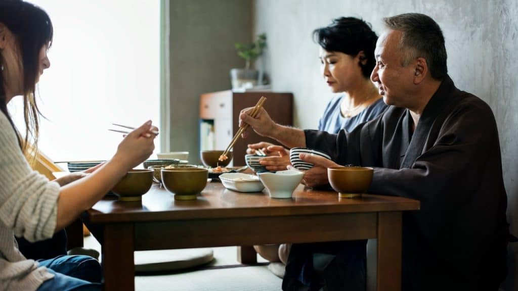 Three people eating a meal with chopsticks at a low wooden table indoors.