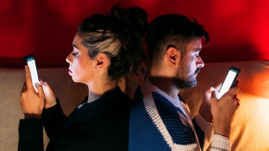 Man and woman sitting back-to-back, absorbed in their smartphones in a dark room.