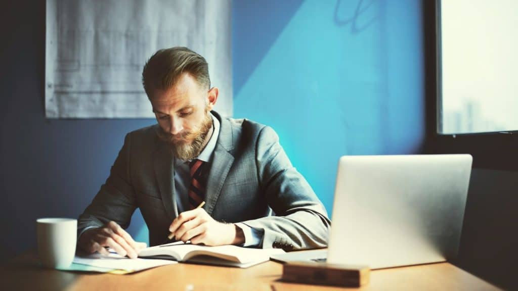 Bearded man in a suit writing in a notebook at a desk with a laptop.