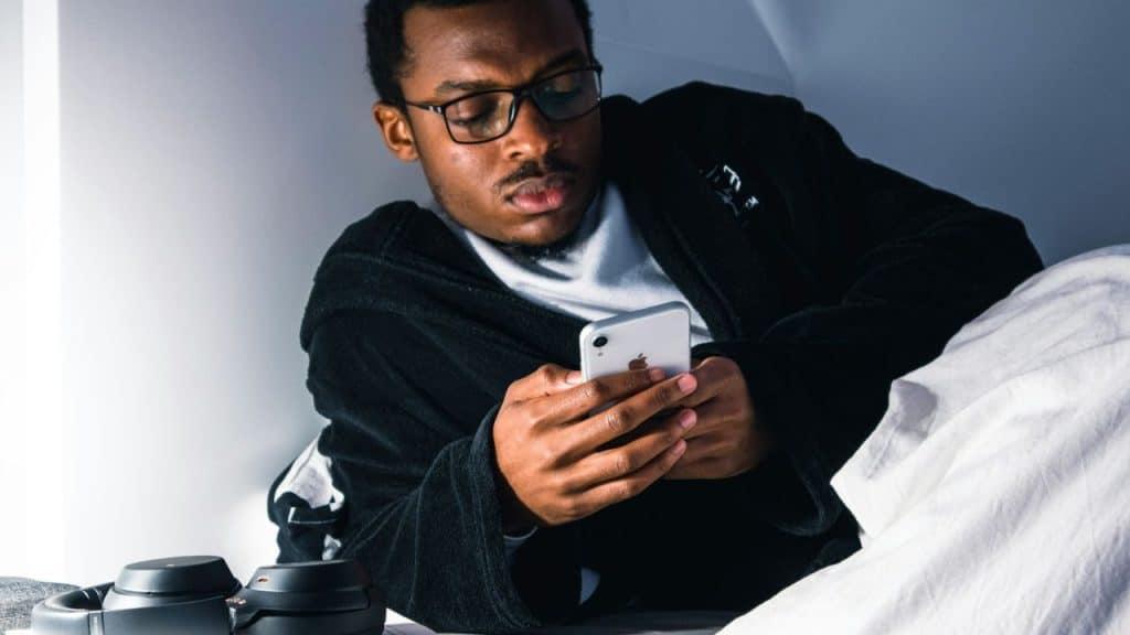 Young man in glasses and a robe lying in bed, intensely focused on his smartphone.