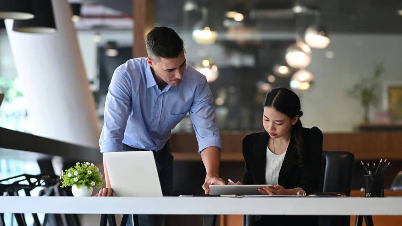 Man leaning over a desk, looking at a tablet held by a seated woman in an office.