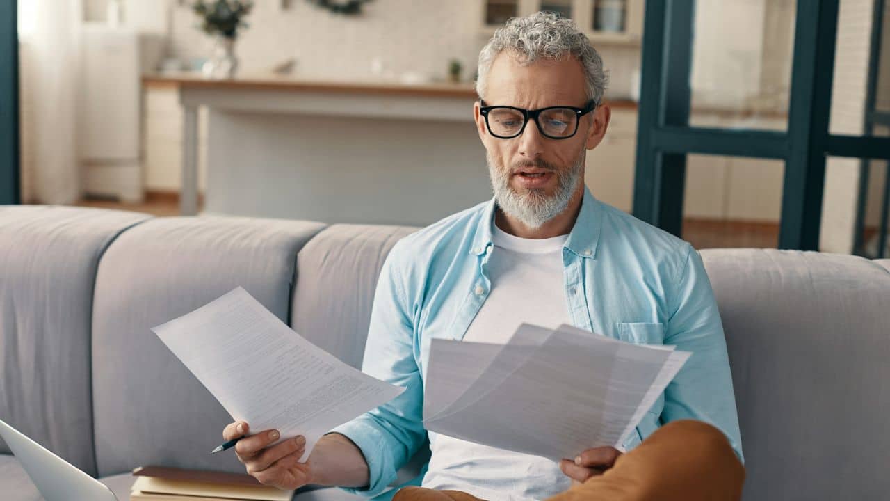 Middle-aged man in a blue shirt and glasses reading documents on a couch.