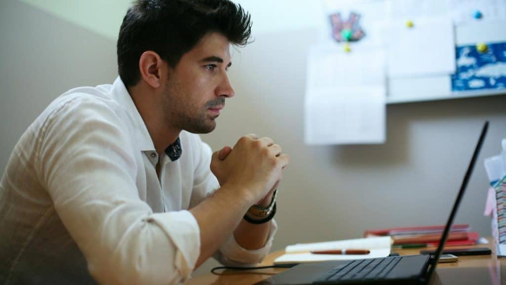 A man in a white shirt sits at a desk, focused on his laptop with hands clasped.
