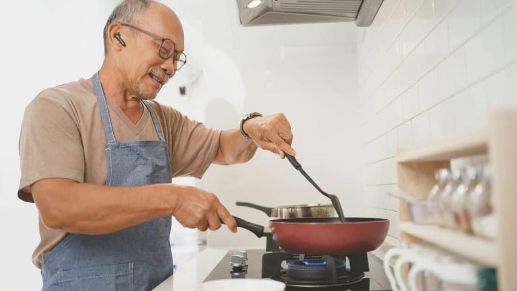 Smiling older man in an apron and glasses cooking at a stove in a kitchen.
