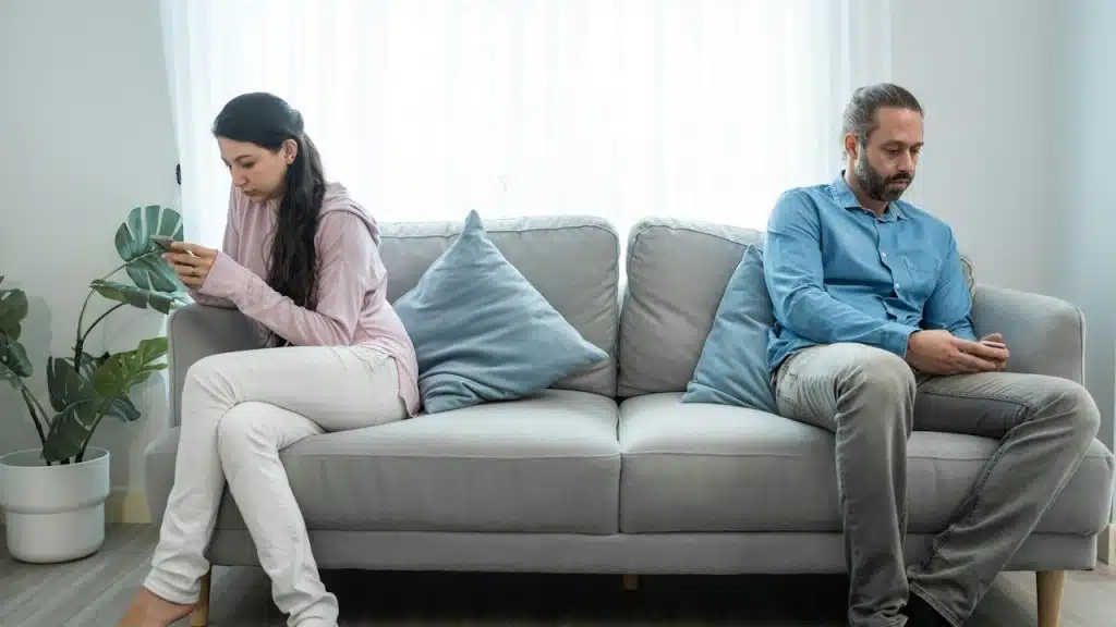 A couple sitting apart from each other on a couch and using their phones.