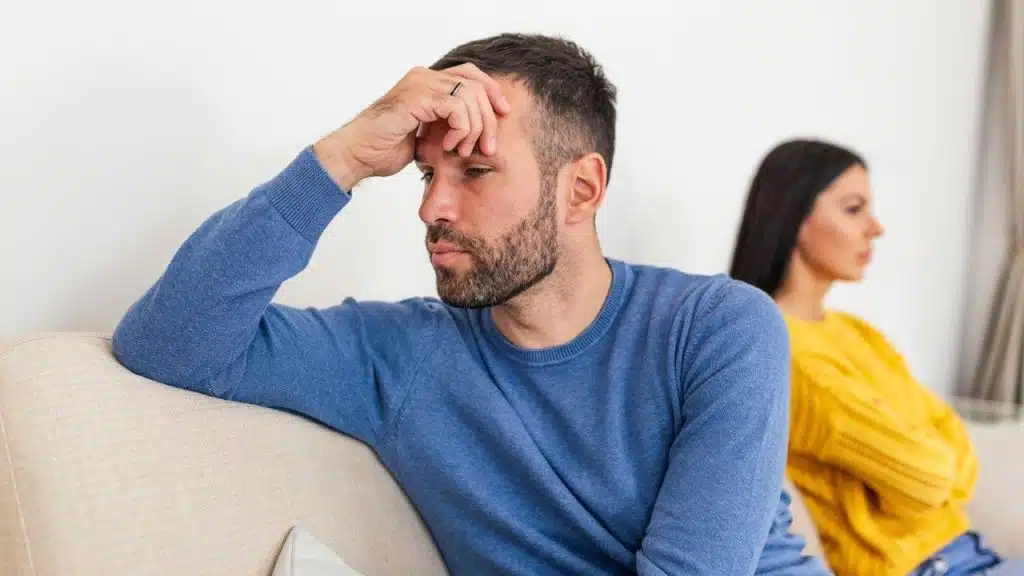 Worried woman looks at stressed man holding his head while sitting on a couch.