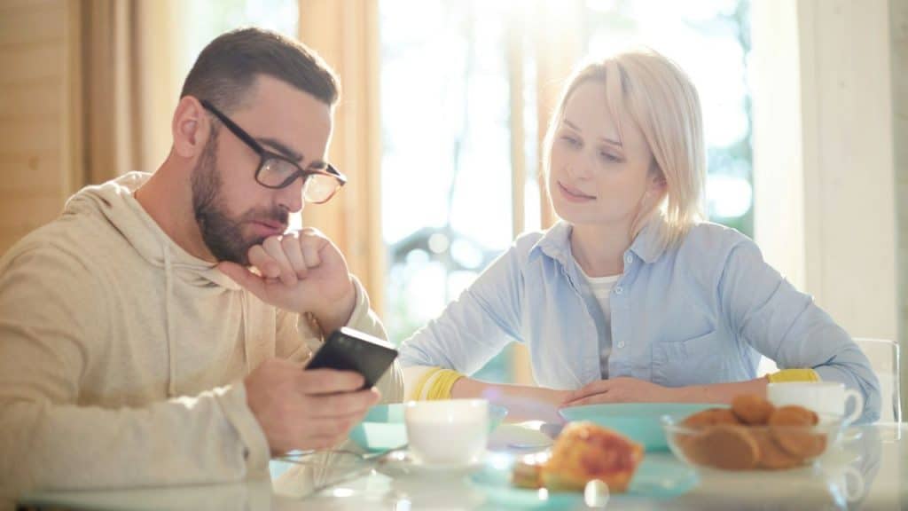 A man looking at his phone at a table while a woman looks at him with a gentle smile.
