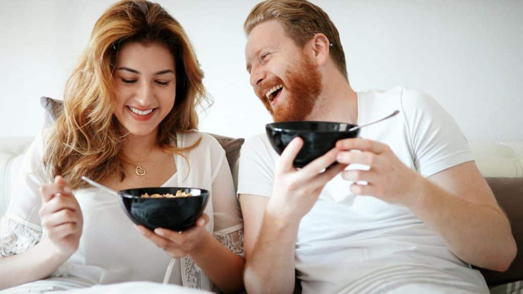 A laughing man and smiling woman eating cereal from black bowls.
