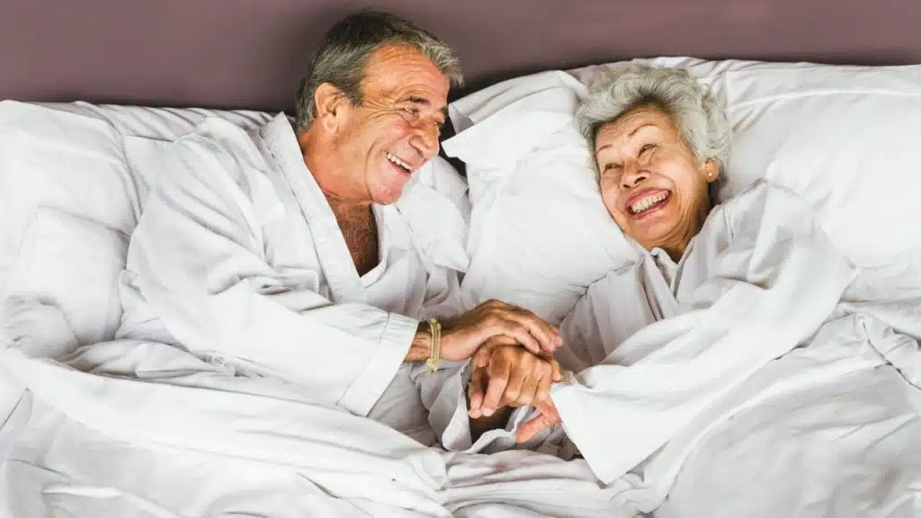An older man and woman in white robes lie in bed, smiling and holding hands.