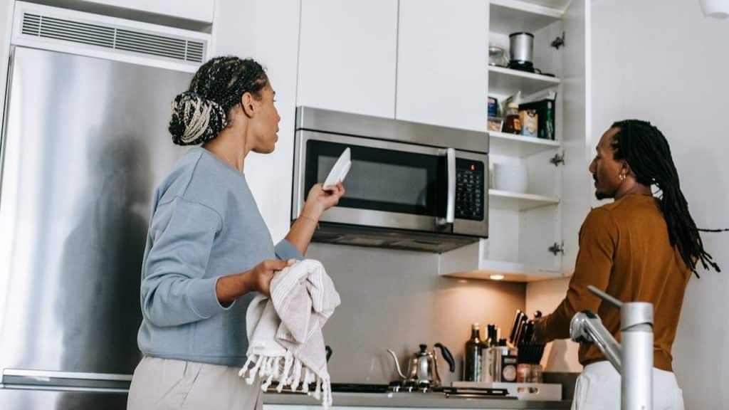 A man and woman standing in a modern white kitchen, looking into cabinets and at the microwave.