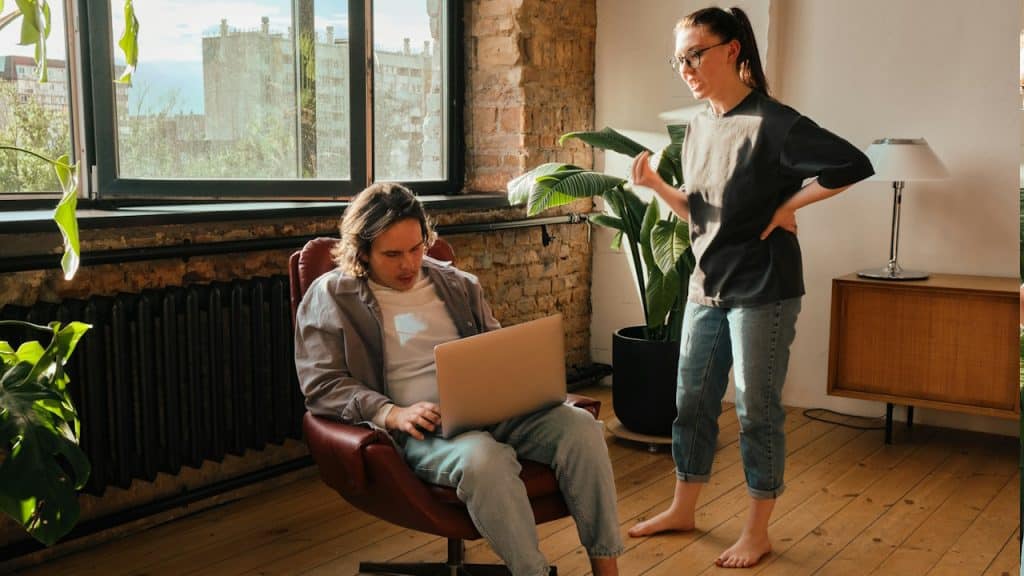 A woman looking at a man as he works on his laptop while sitting in a chair in their living room.