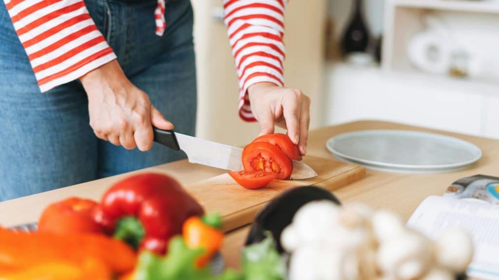 A person slicing a tomato on a wooden cutting board in a kitchen.