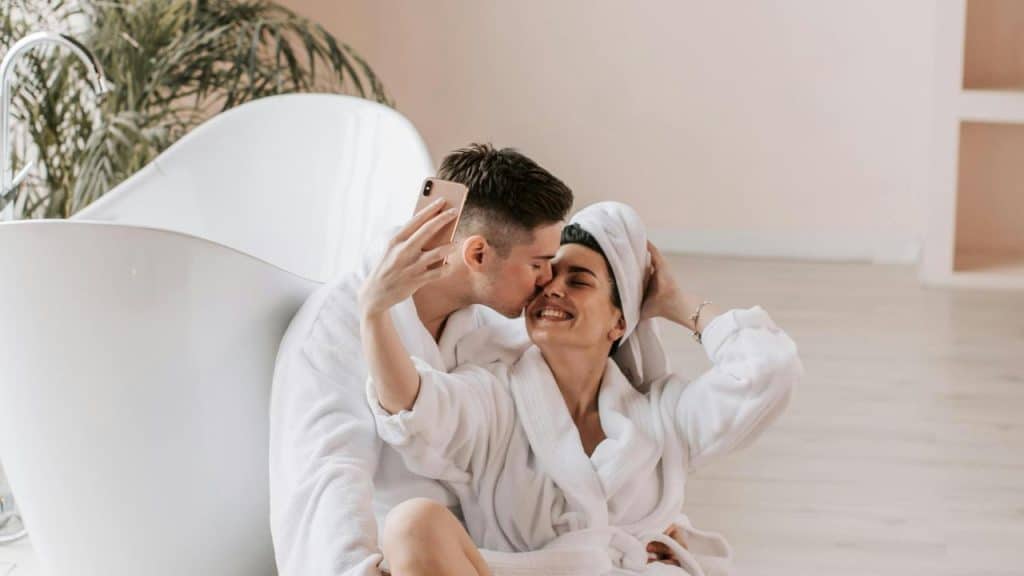 A couple in bathrobes taking a selfie while sitting by a bathtub.