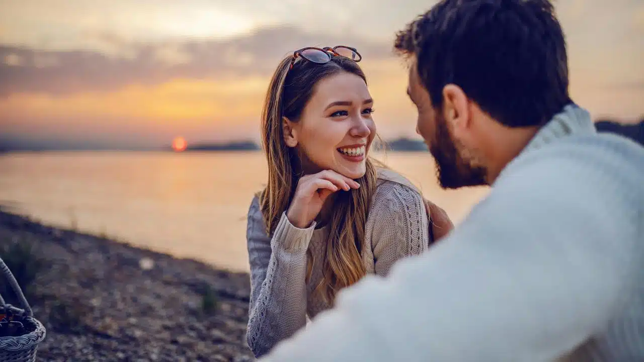 A smiling couple sitting by the water at sunset, looking at each other.