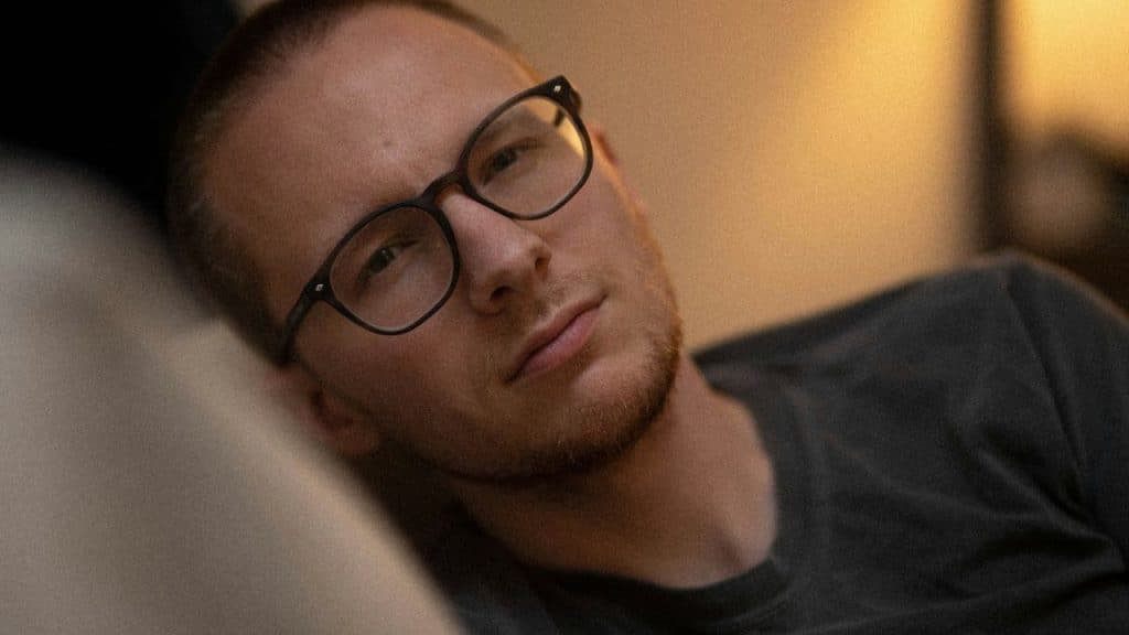 A man with a shaved head and black-rimmed glasses is resting, looking intently at the camera.