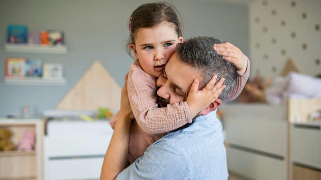 A bearded man holds and hugs a young girl with a pink headband in a child's room.