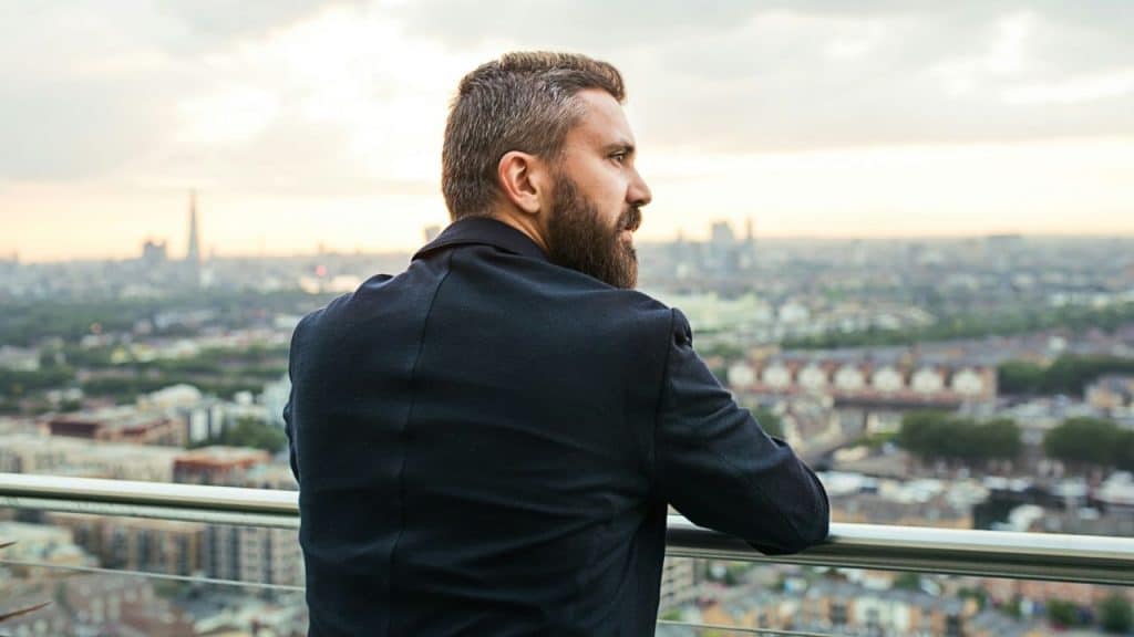 A bearded man in a suit jacket looks out over a cityscape from a rooftop.