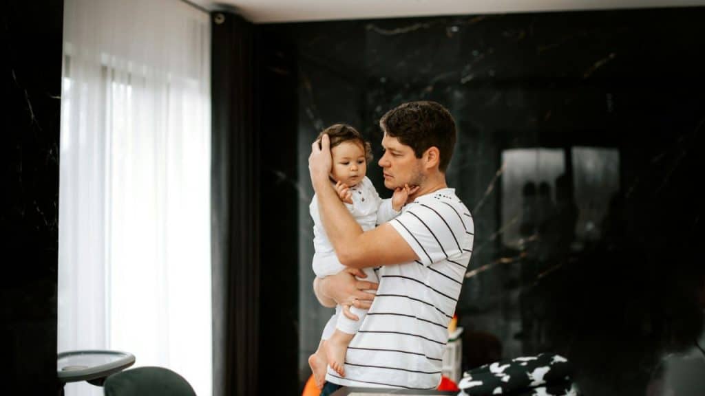 A man in a striped shirt holds and looks at a baby with dark hair.