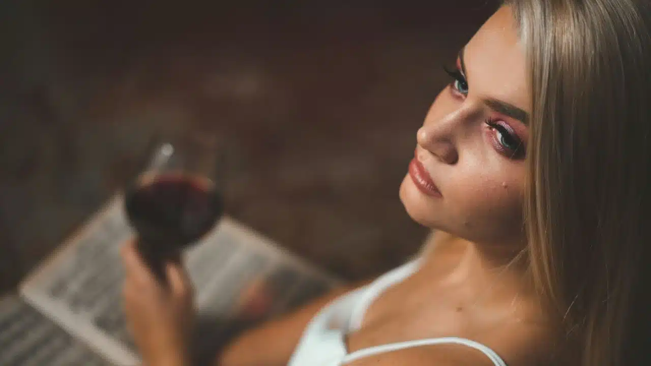A blonde woman holds a glass of red wine, looking up over her shoulder.