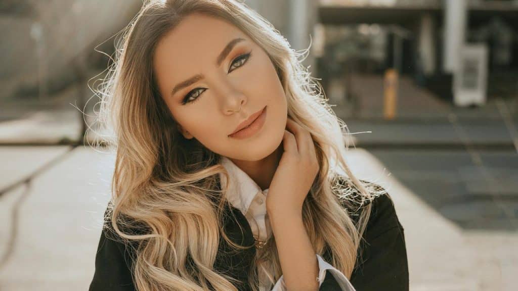 A woman with long blonde hair posing outdoors with her hand near her neck.