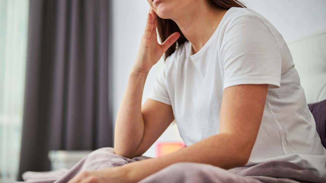 A woman sitting on a bed touching her jaw with a pained expression.