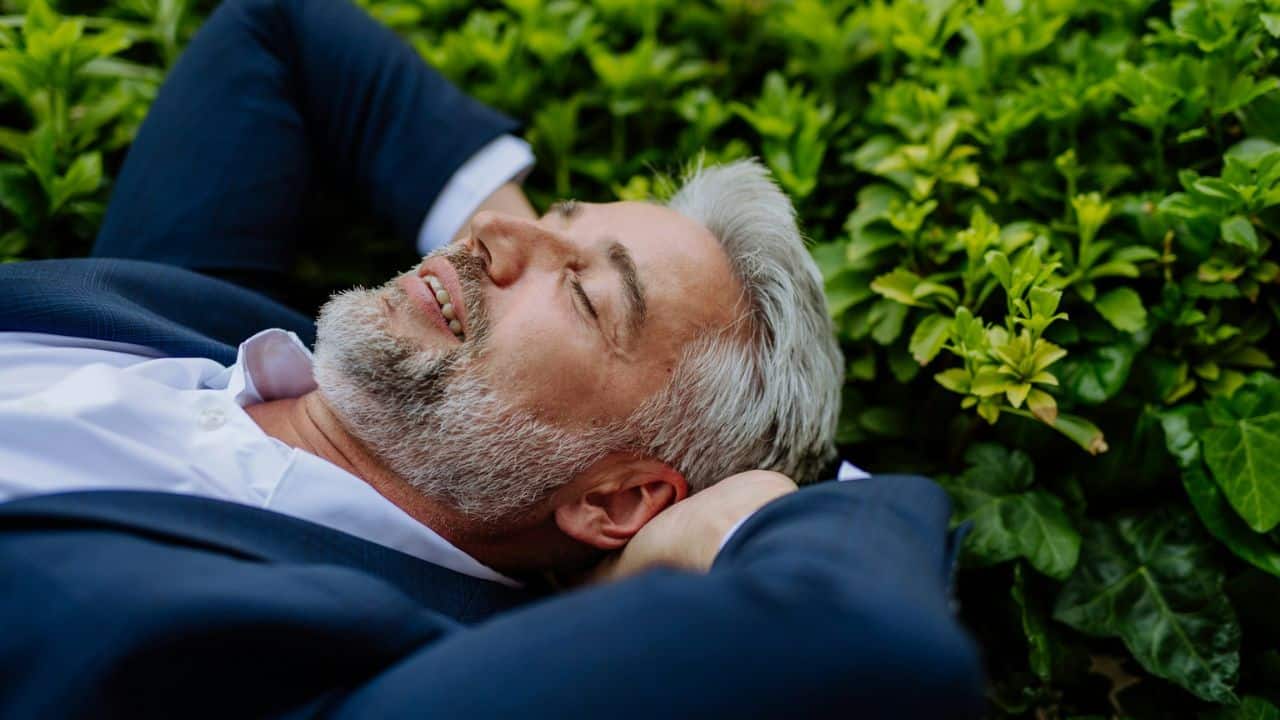 A smiling man with gray hair is lying in green foliage with his eyes closed.