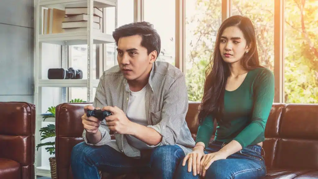 A man is intensely focused on playing a video game with a controller while a woman sits next to him looking annoyed.