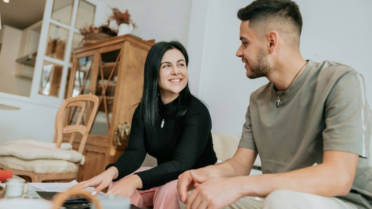 A smiling woman and a man are talking while sitting on a couch in a living room.