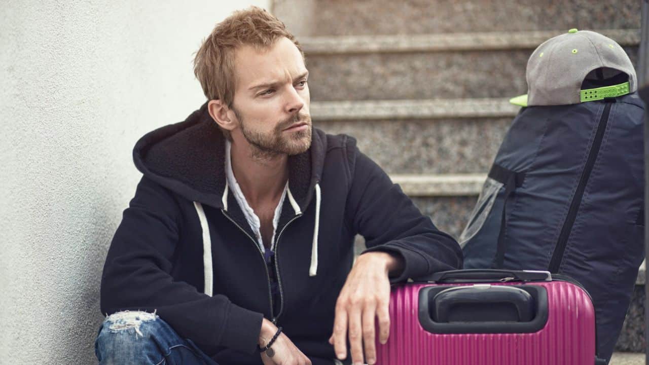 A sad man in a black hoodie is sitting on steps with a bright pink suitcase.
